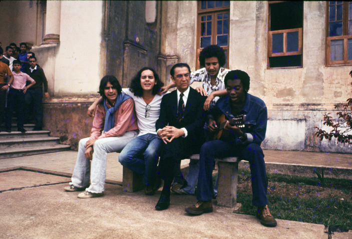 Part of the Clube da Esquina, sitting on a bench: Lô Borges, Fernando Brant, Márcio Borges, and Milton Nascimento. Ex-president of Brazil Juscelino Kubitscheck is also there, looking very serious in a black suit and tie