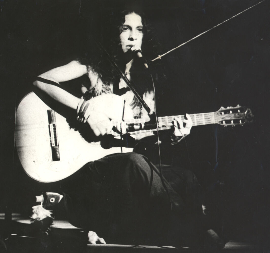 Gal Costa sitting down, playing guitar and singing